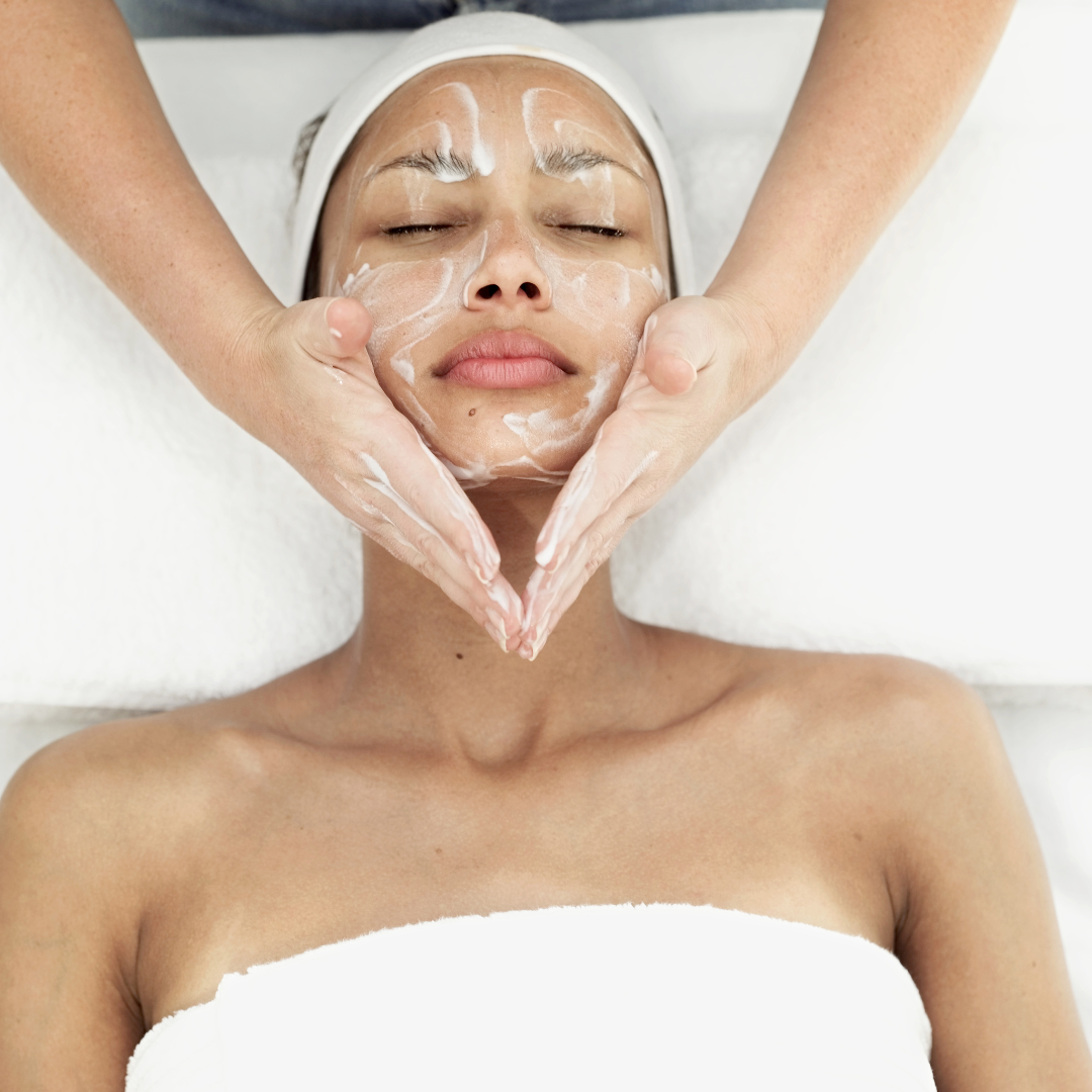 A person receiving a facial massage with white cream applied to their face, lying down in a spa setting.