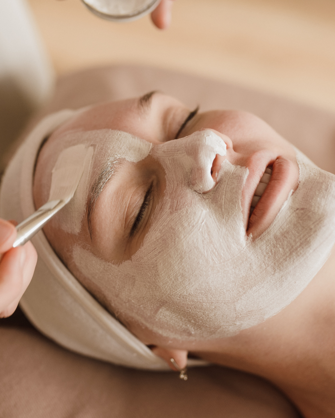 A person receiving a clay facial mask application, with a tool being used to smooth the product onto their forehead.