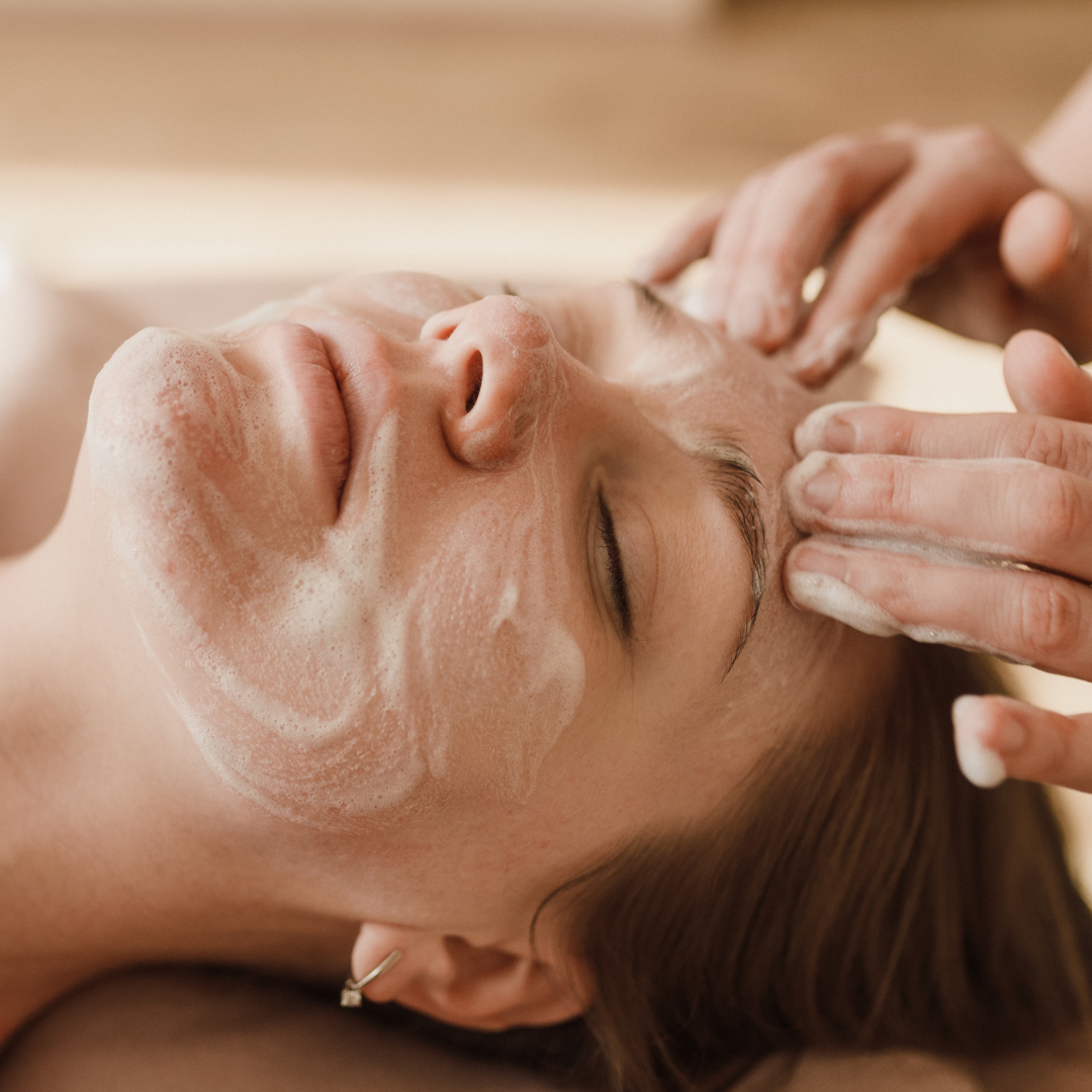 A person lying down receiving a facial treatment with cleanser applied to their face as hands gently massage their forehead.