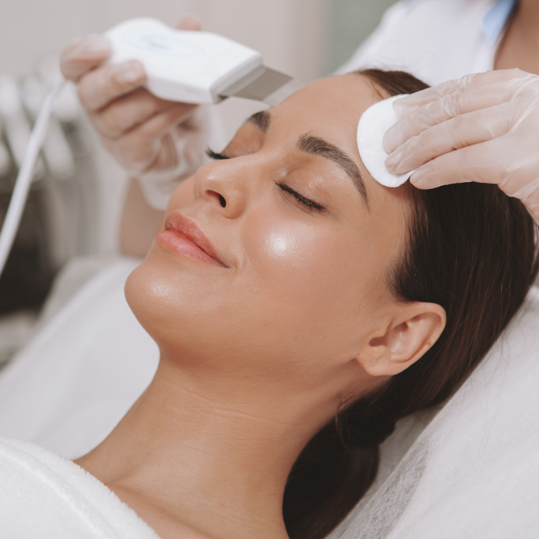 A person lying on a spa bed receives a professional facial treatment using an ultrasonic skin spatula and a cotton pad.