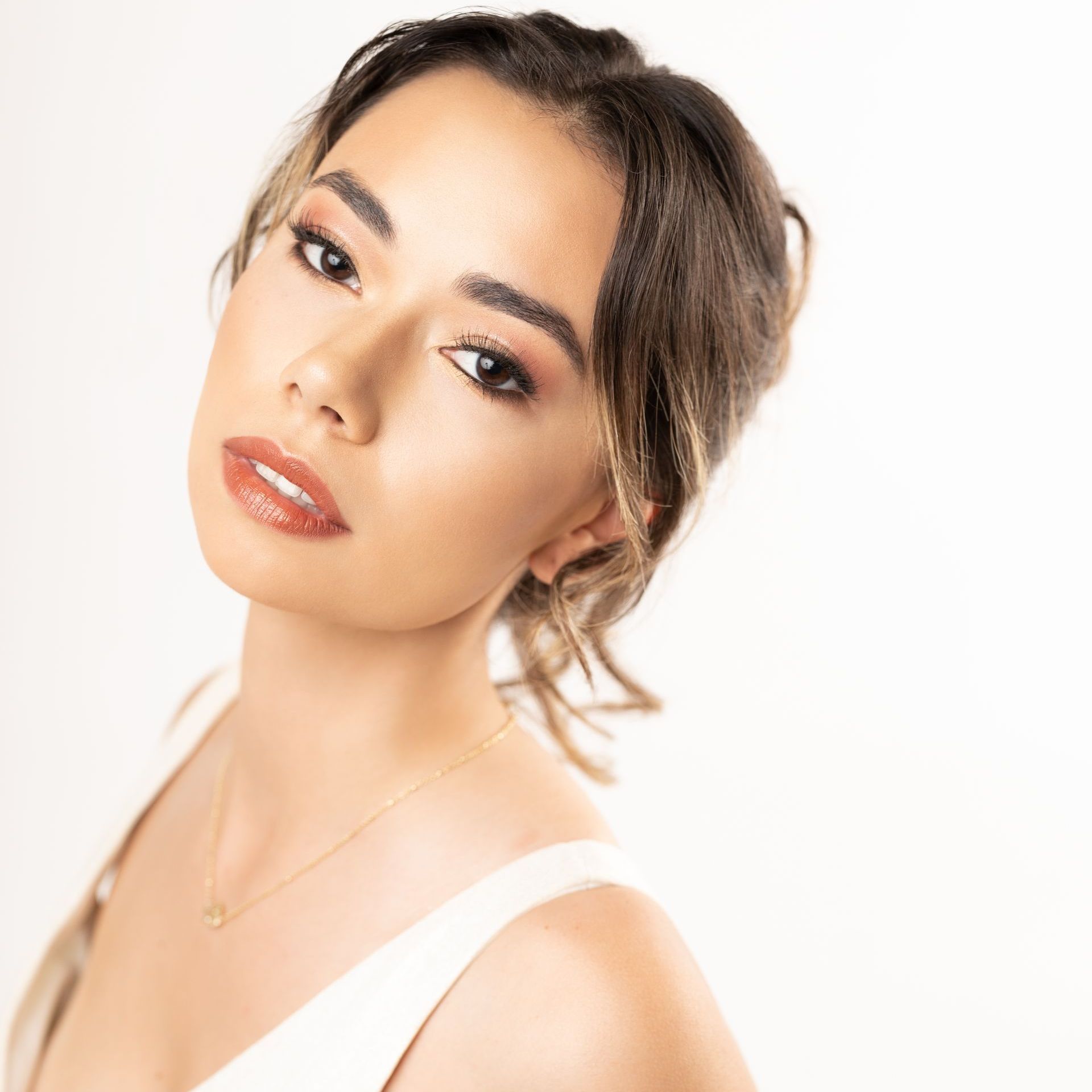 A headshot of a person with dark hair, light makeup, and a white top, gazing directly into the camera against a white wall.