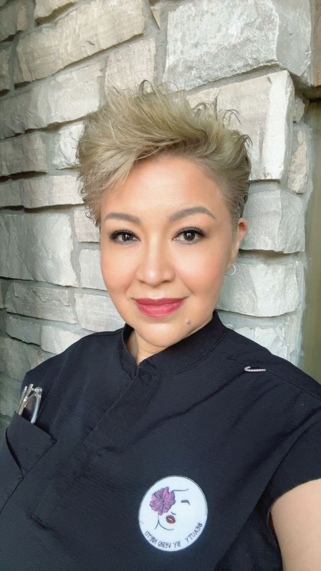 A person with short, blonde hair and a dark shirt poses in front of a stone wall. They have a small logo on their chest.