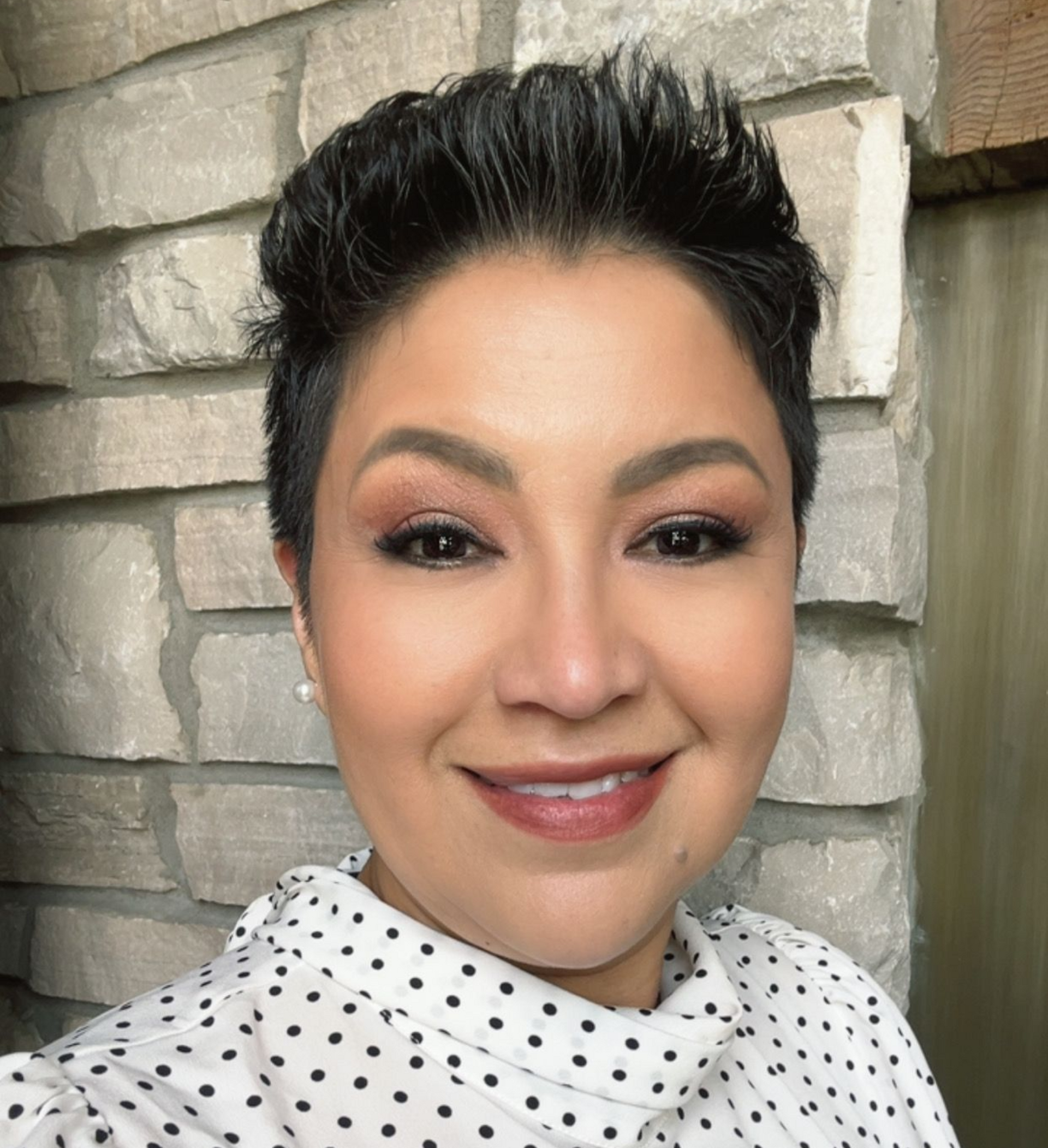 A person with a short, dark pixie cut smiles at the camera, wearing a white polka-dot blouse against a light stone wall.