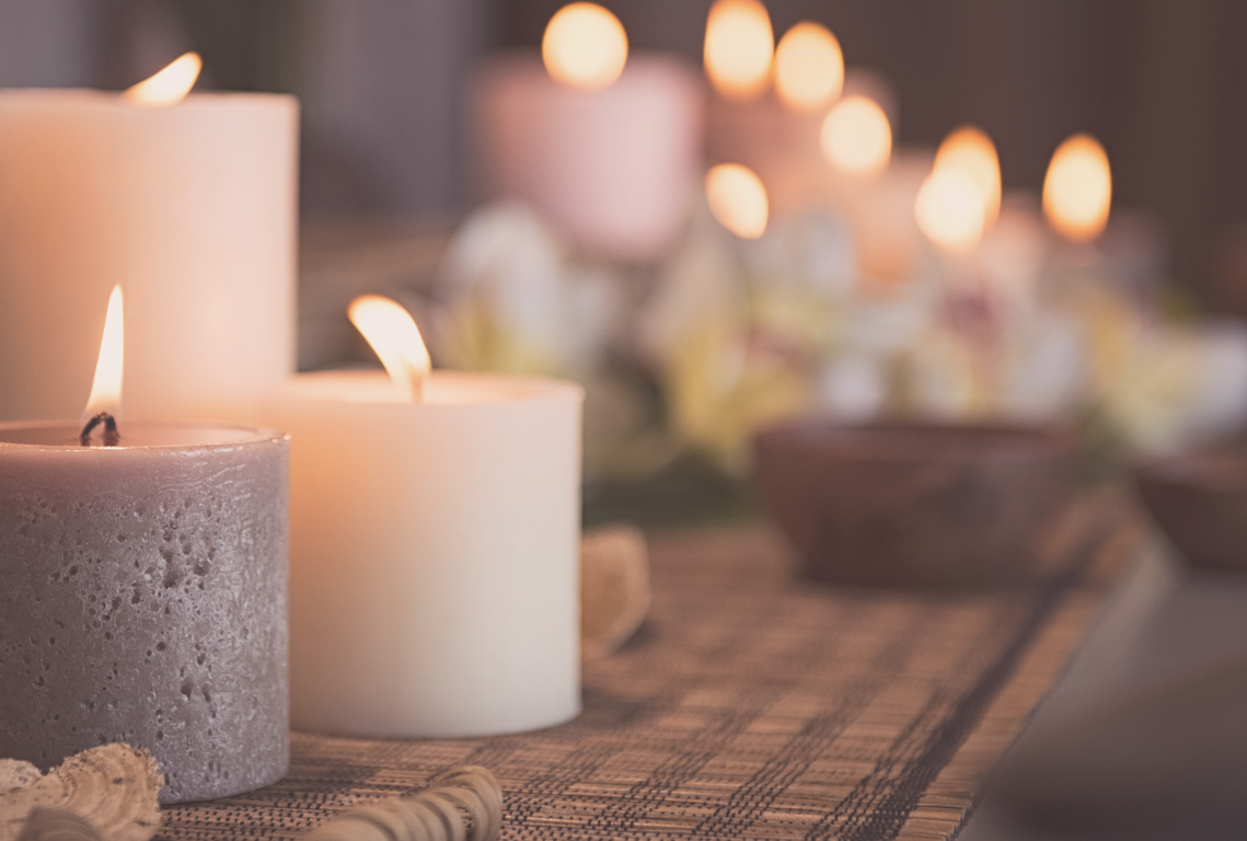 Softly lit candles in varying sizes and shades of white and grey glow against a warm, textured tabletop surface.