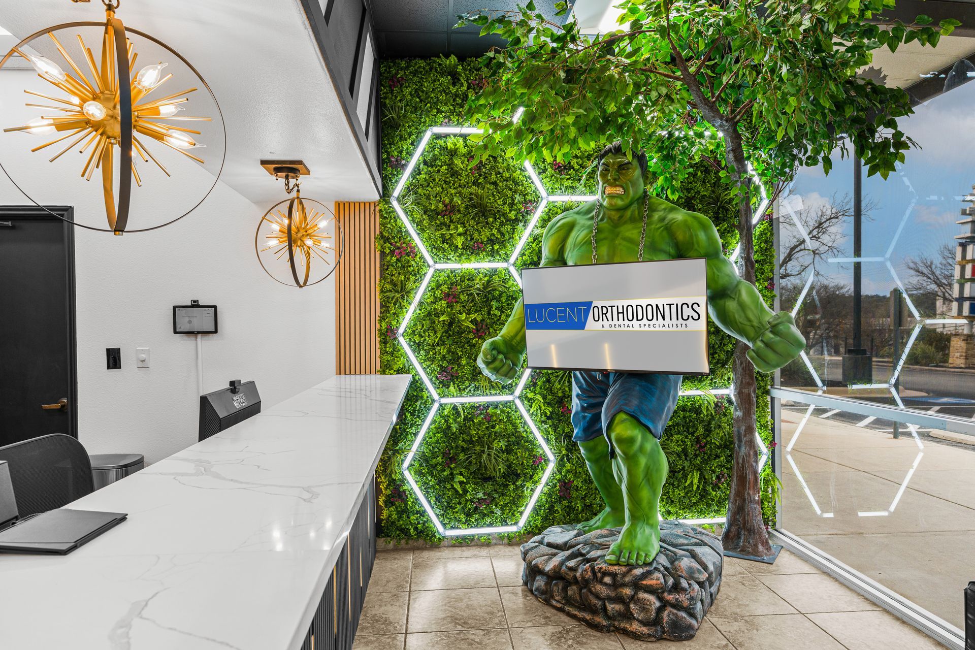 Lucent Orthodontics, San Antonio. A life-sized Hulk statue stands in a modern office reception area in front of a green plant wall with neon hexagon lights.