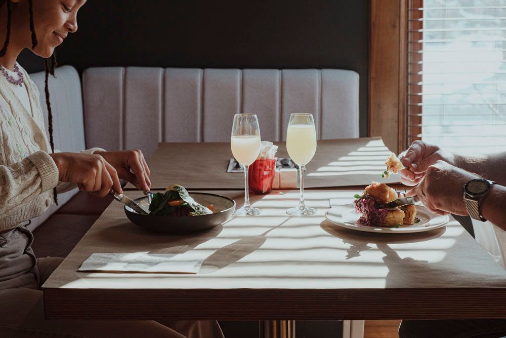 Two people dining in a sunlit booth with plates of food, drinks, and a white tablecloth.