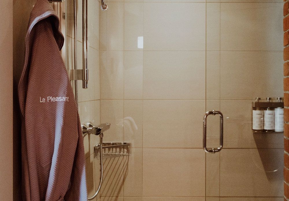Un peignoir avec le logo de Le Pleasant est suspendu dans une salle de bain à côté d'une douche.