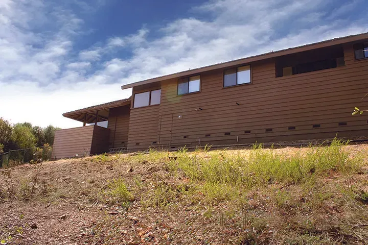 Brown-sided house on a hillside under a cloudy blue sky. Sparse grass in foreground.