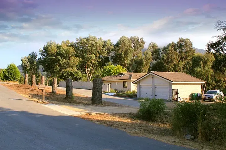 A one-story beige house with a two-car garage, surrounded by trees and a roadway.