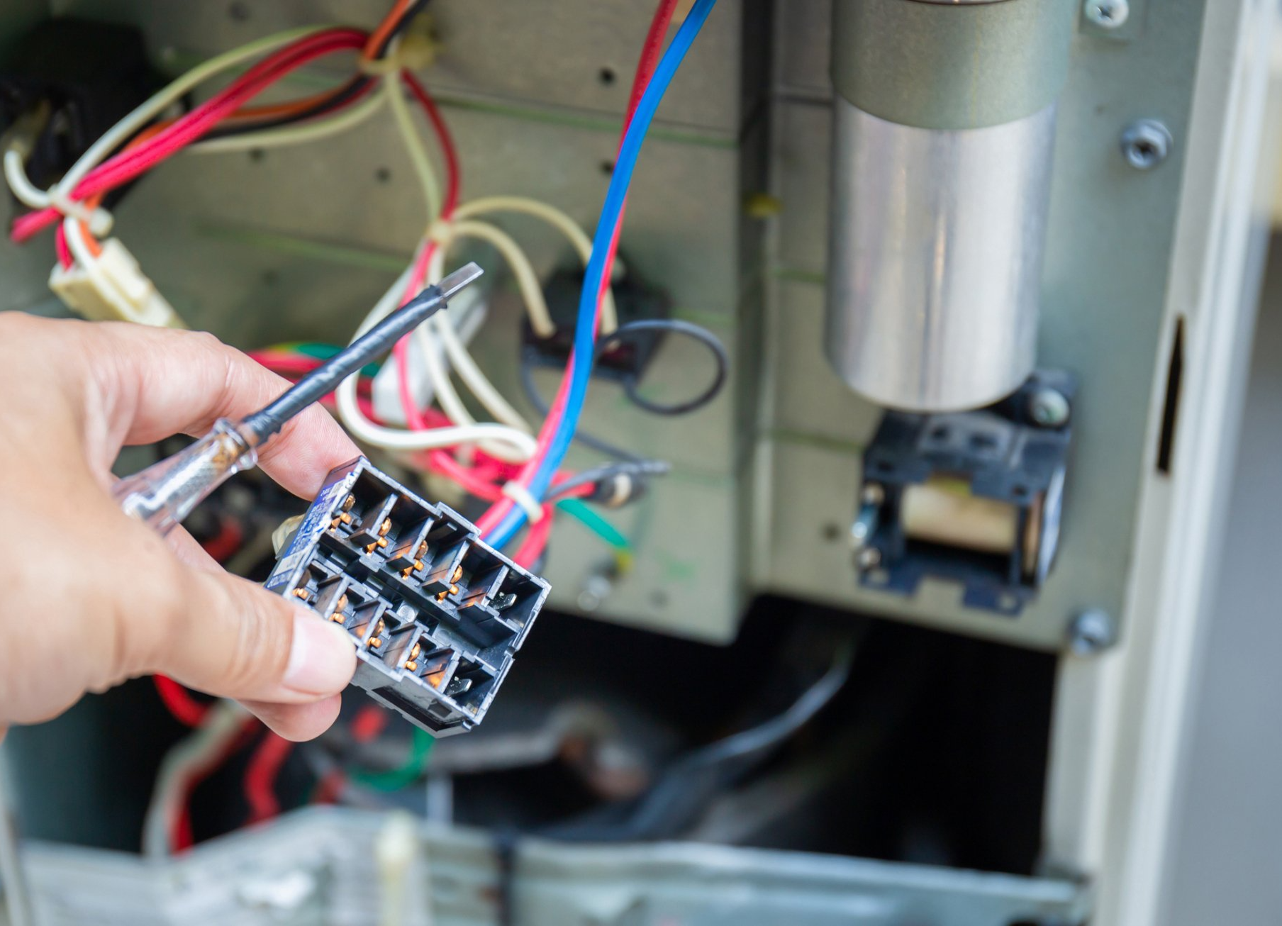 Person holding electrical connector with screwdriver, repairing appliance. Wires, capacitor, and interior parts visible.
