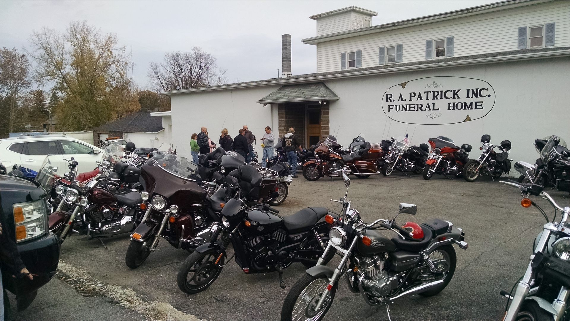 Motorcycles parked outside R. Patrick Inc. Funeral Home; a group of people stand nearby on an overcast day.