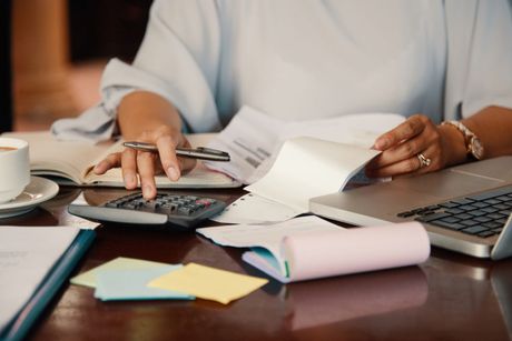 Woman calculating on a desk with papers, a calculator, laptop, coffee cup, and sticky notes.