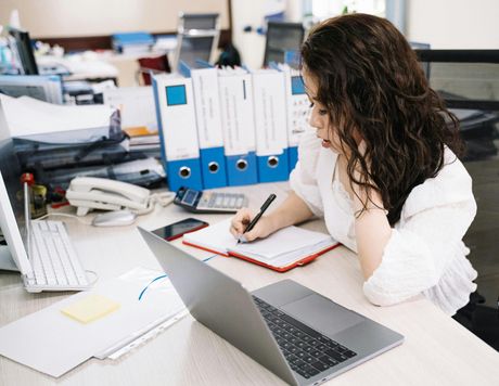 Woman writing in a notebook at a desk with a laptop, calculator, and papers in an office setting.