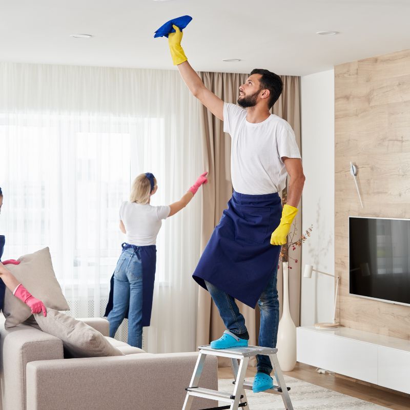 Three people cleaning a living room. Man on a step ladder wipes the ceiling, others dust furniture.