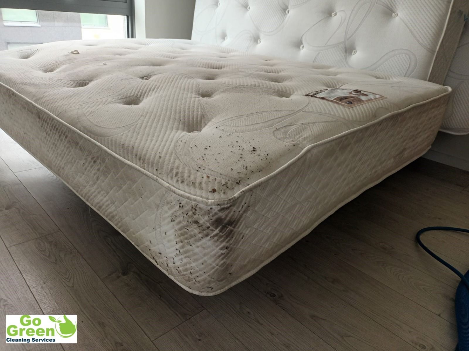 A stained, soiled mattress sits on a wood floor near a wall with a window.