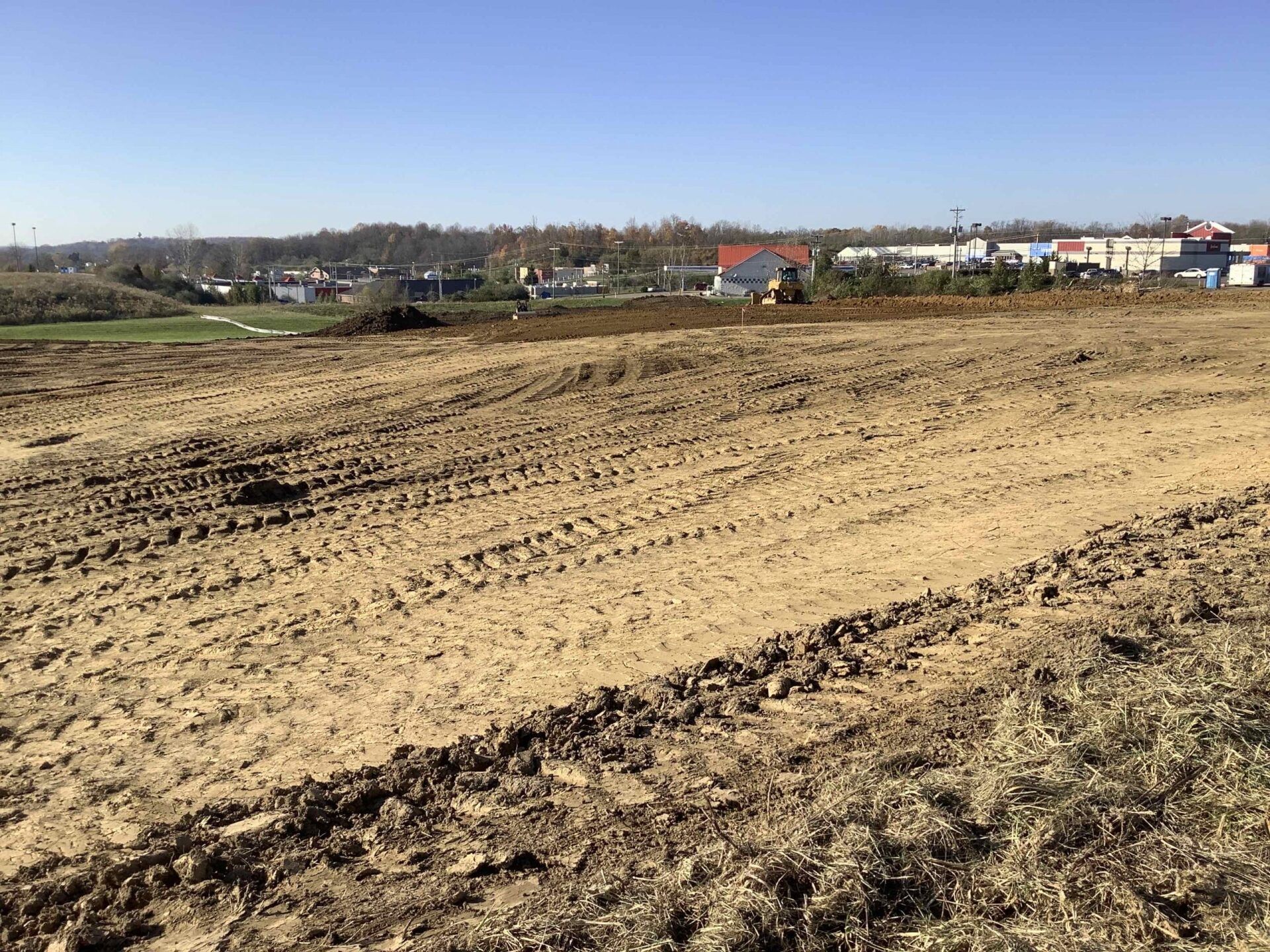 Dirt field with tire tracks; buildings in the distance under a blue sky.