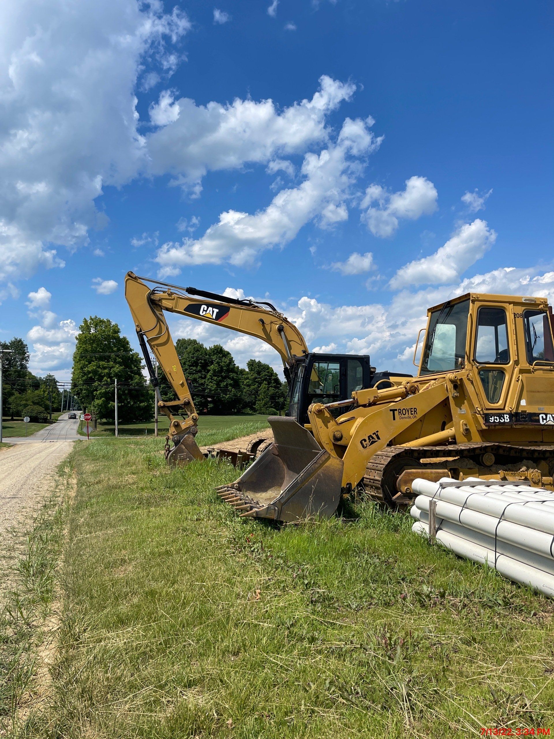 Yellow CAT excavator on a grassy area, digging near white drainage pipes, under a blue sky with clouds.