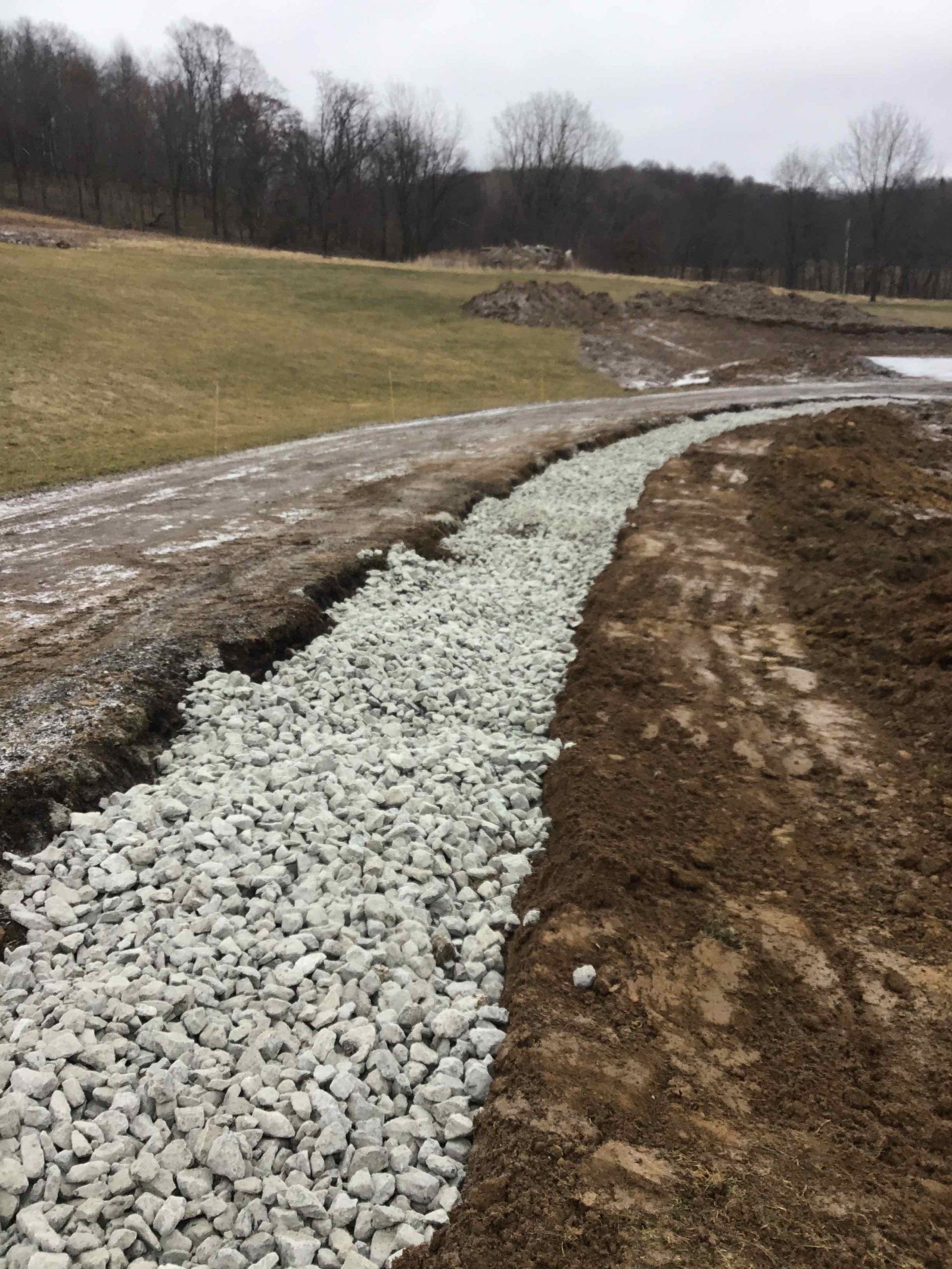 Ditch filled with gravel along a dirt path, next to a grassy area and trees.