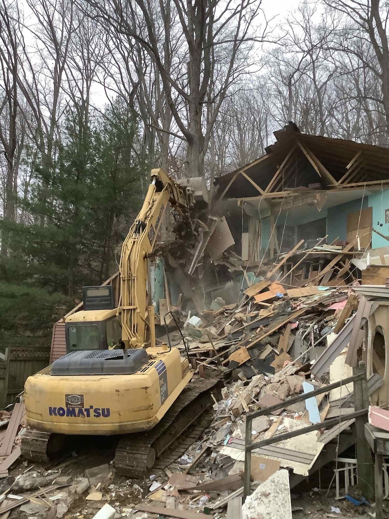 Yellow excavator demolishing a house, debris flying. Trees in background. Cloudy day.