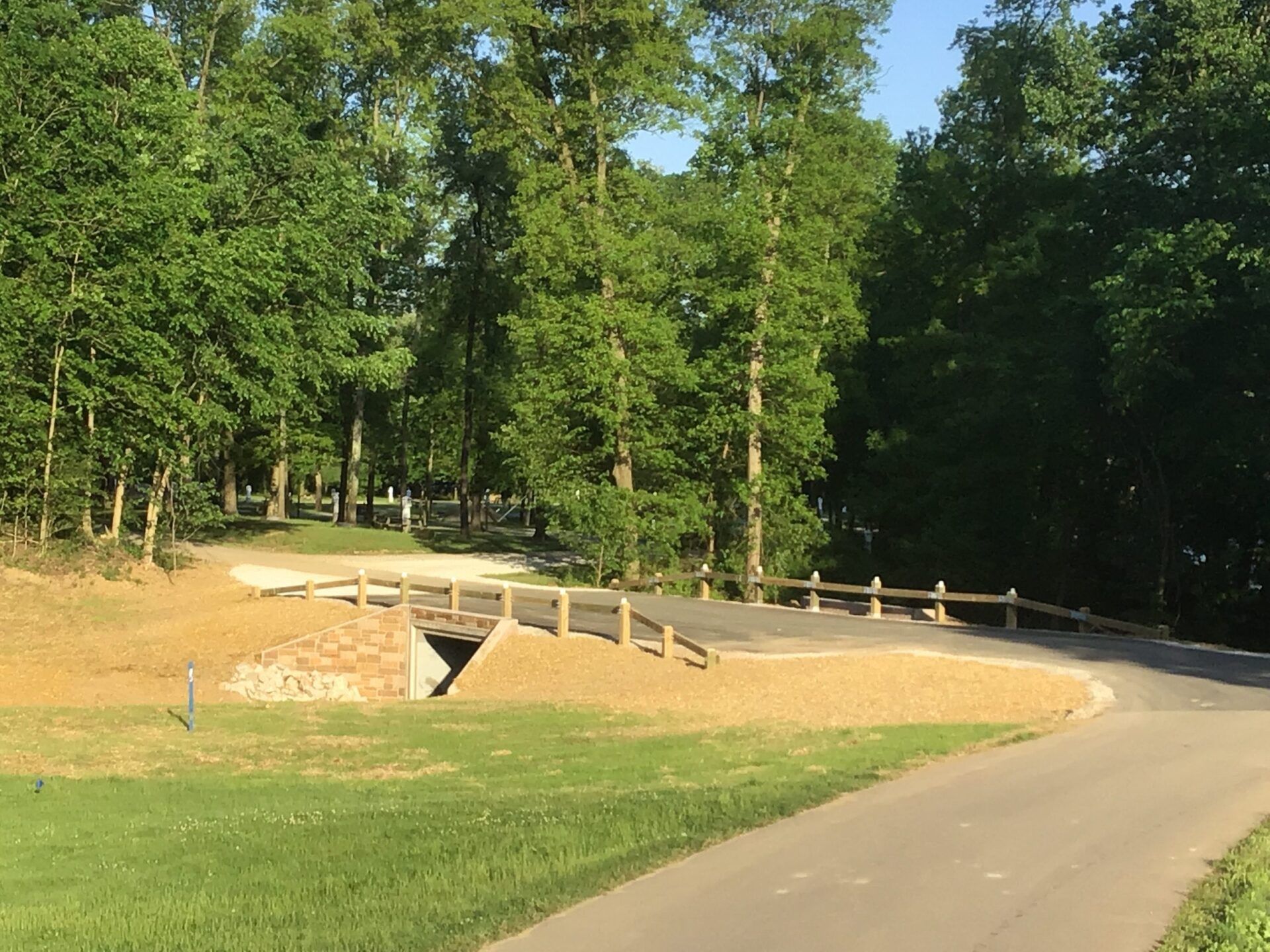 Bridge over a small tunnel in a park, flanked by trees and a paved path.