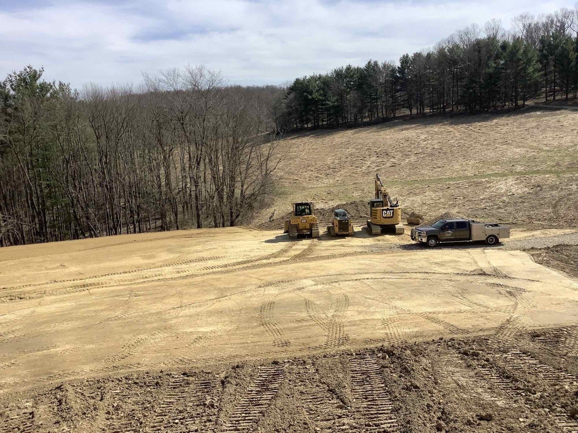 Construction site with bulldozers and a truck clearing land in a hilly, wooded area.