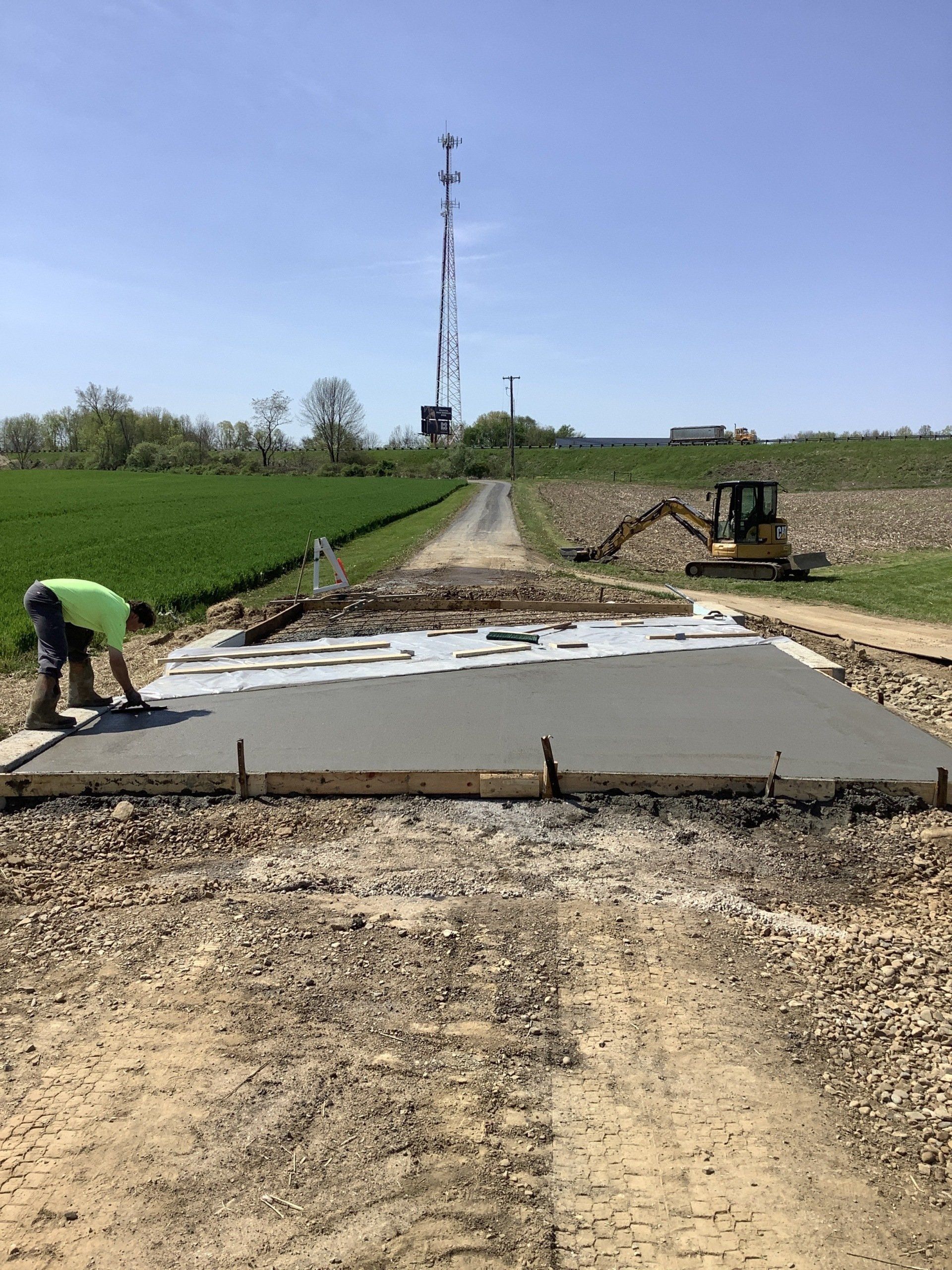 Man pours concrete on a construction site; gravel road, tall tower in background.