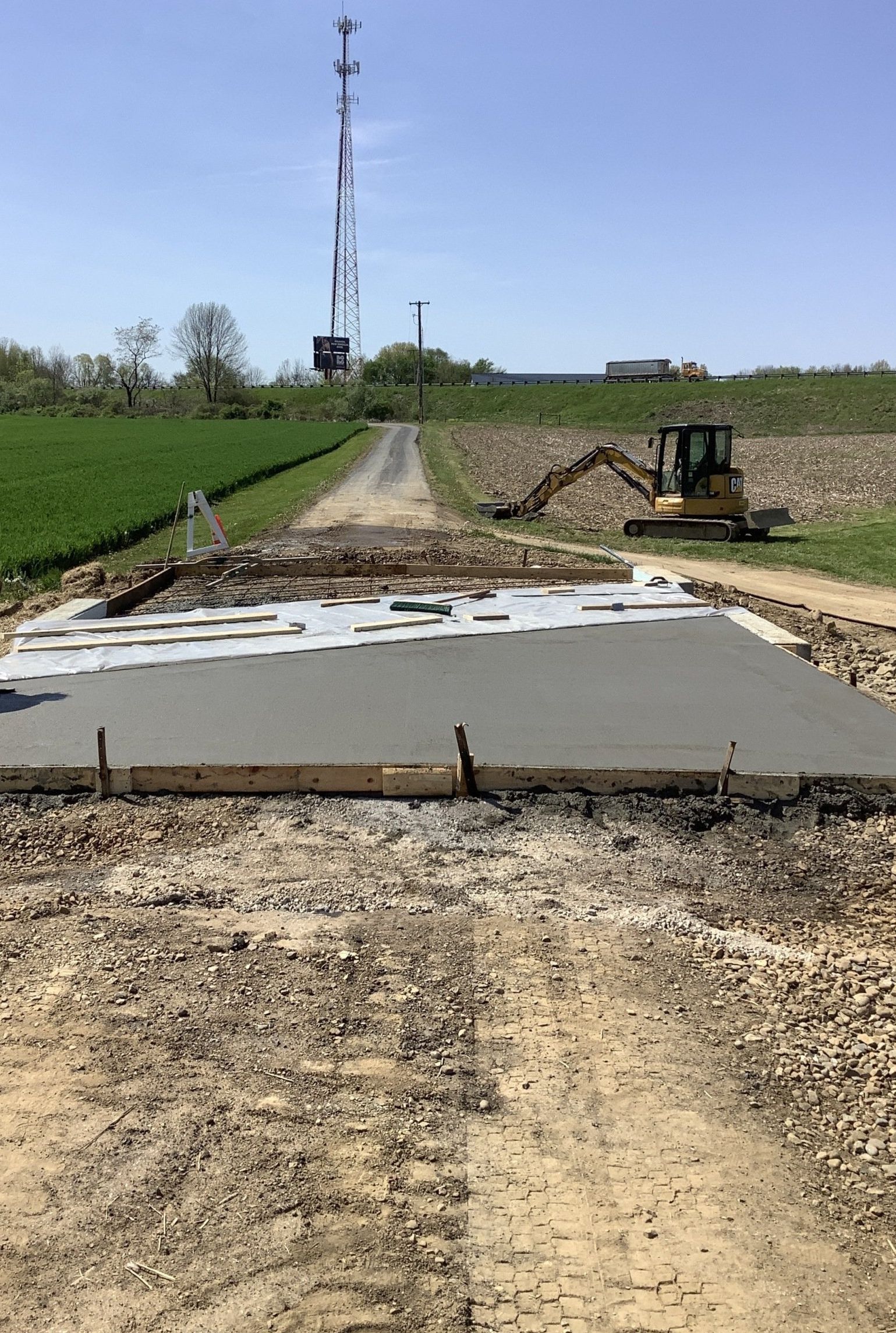 Construction site with newly poured concrete pad in a dirt road, excavator in the background, and radio tower in distance.