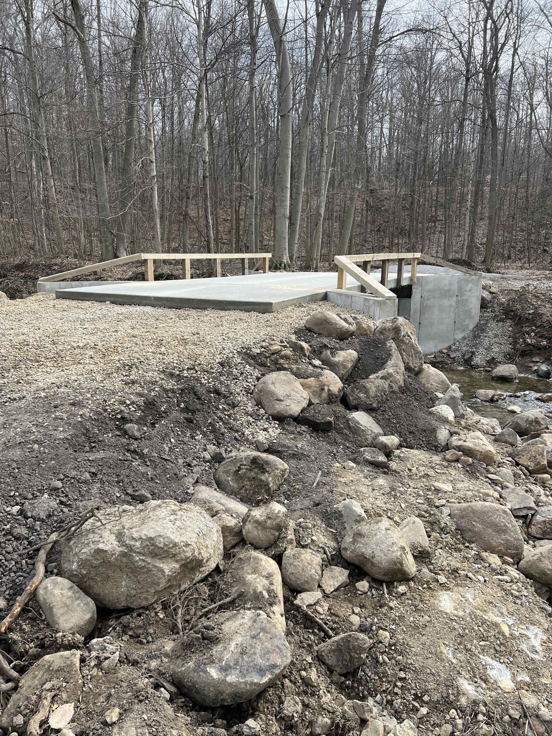 A concrete platform with wooden benches and a retaining wall in a wooded area, surrounded by rocks and dirt.