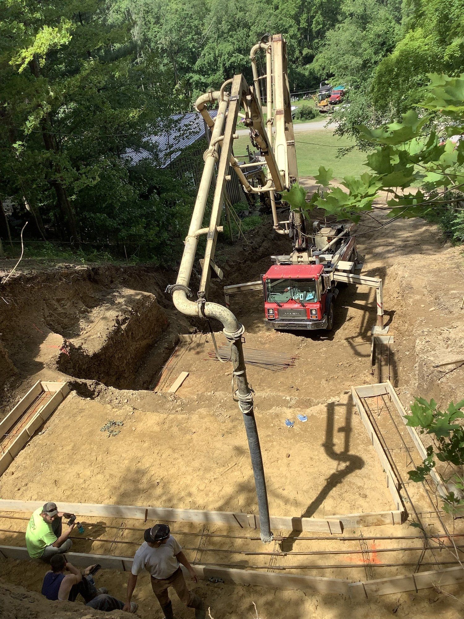 Concrete pump truck pouring cement into a foundation. Two workers in hard hats watch. Green trees frame the scene.