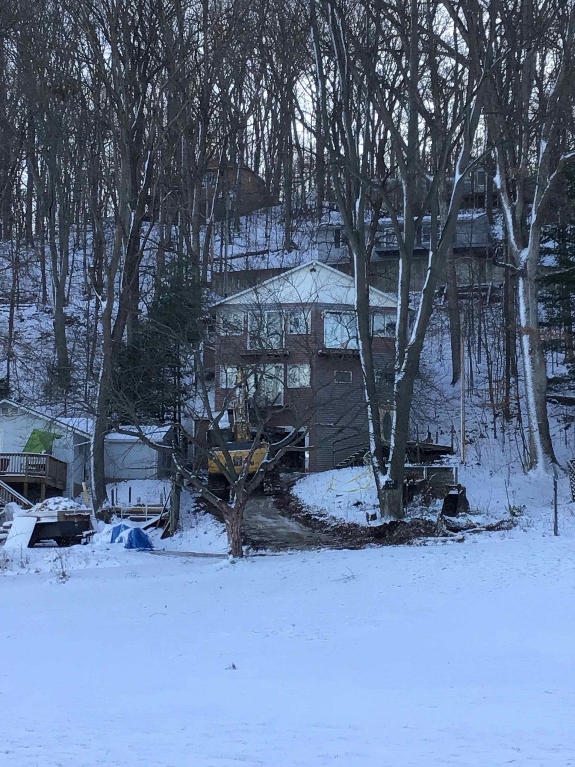 Snowy scene of a multi-story house on a hillside, surrounded by trees. An excavator sits near a path.