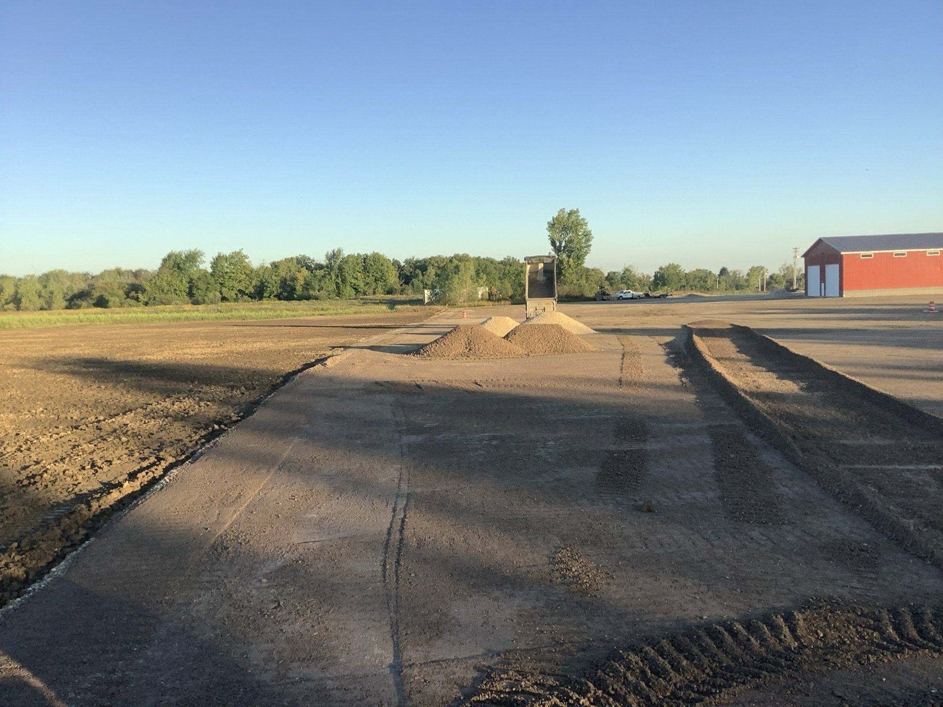 A construction site with a freshly graded gravel surface, soil, and a red building in the background.
