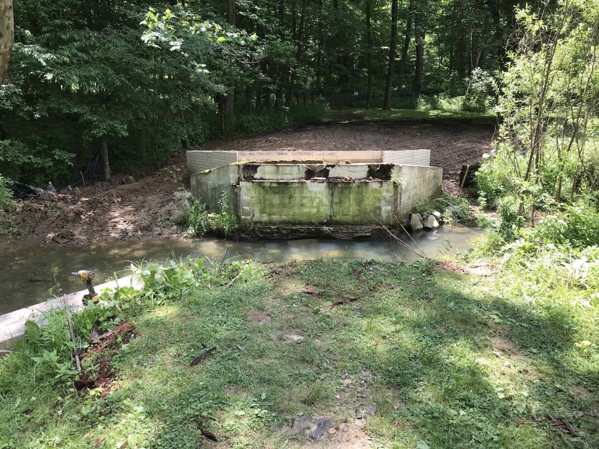 A small, mossy concrete bridge spans a shallow stream in a wooded area.