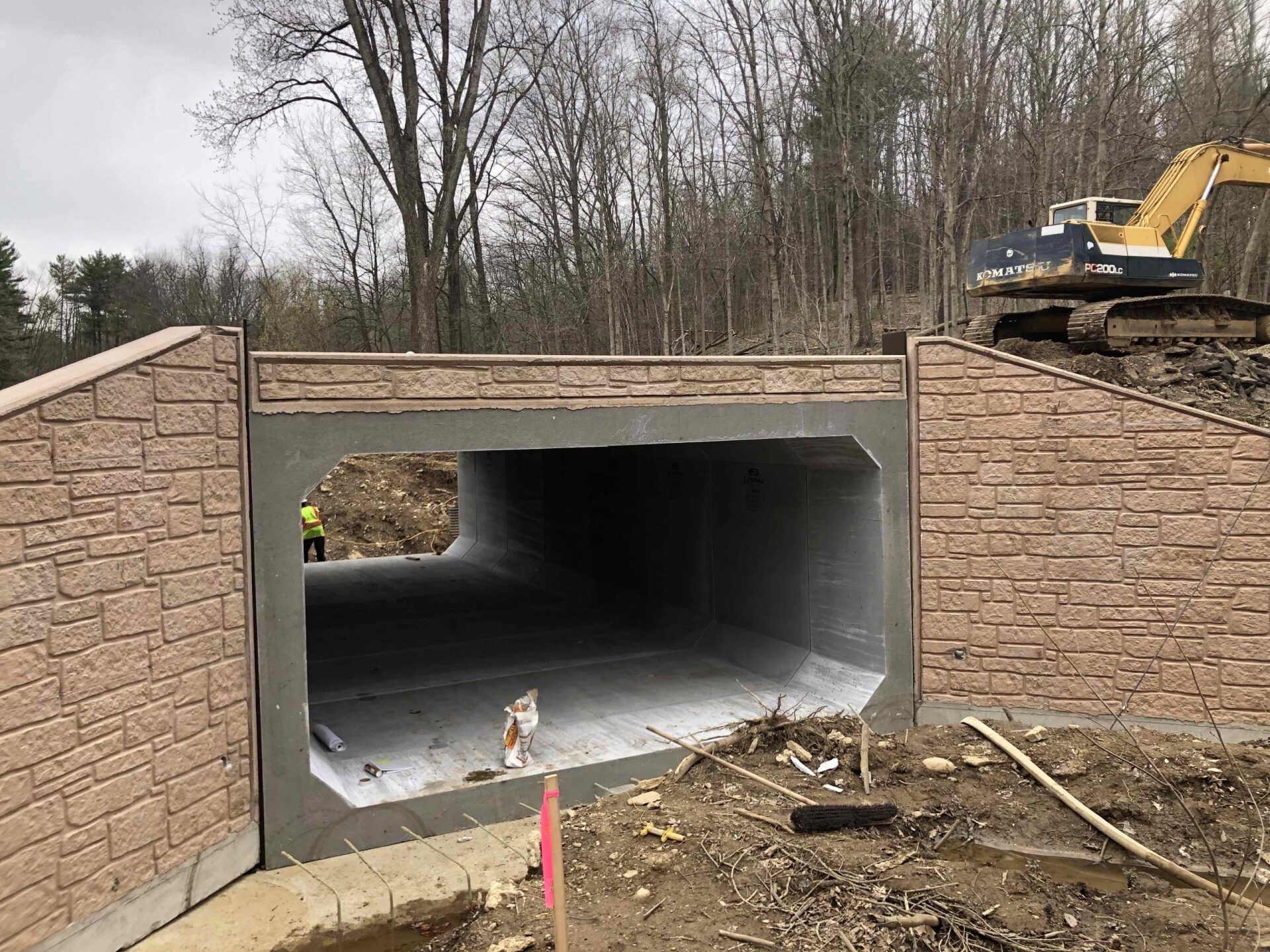 Concrete tunnel under construction, framed by brick walls. An excavator is on the right, trees in background.