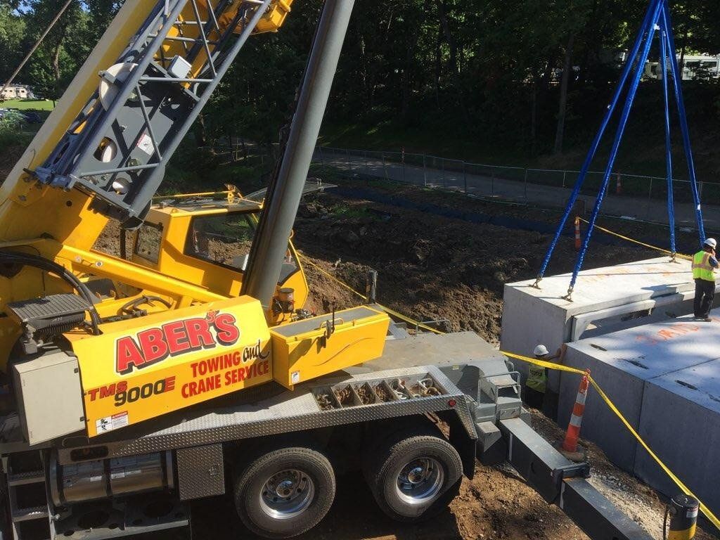 A yellow crane lifts a concrete slab at a construction site. Workers in vests observe.