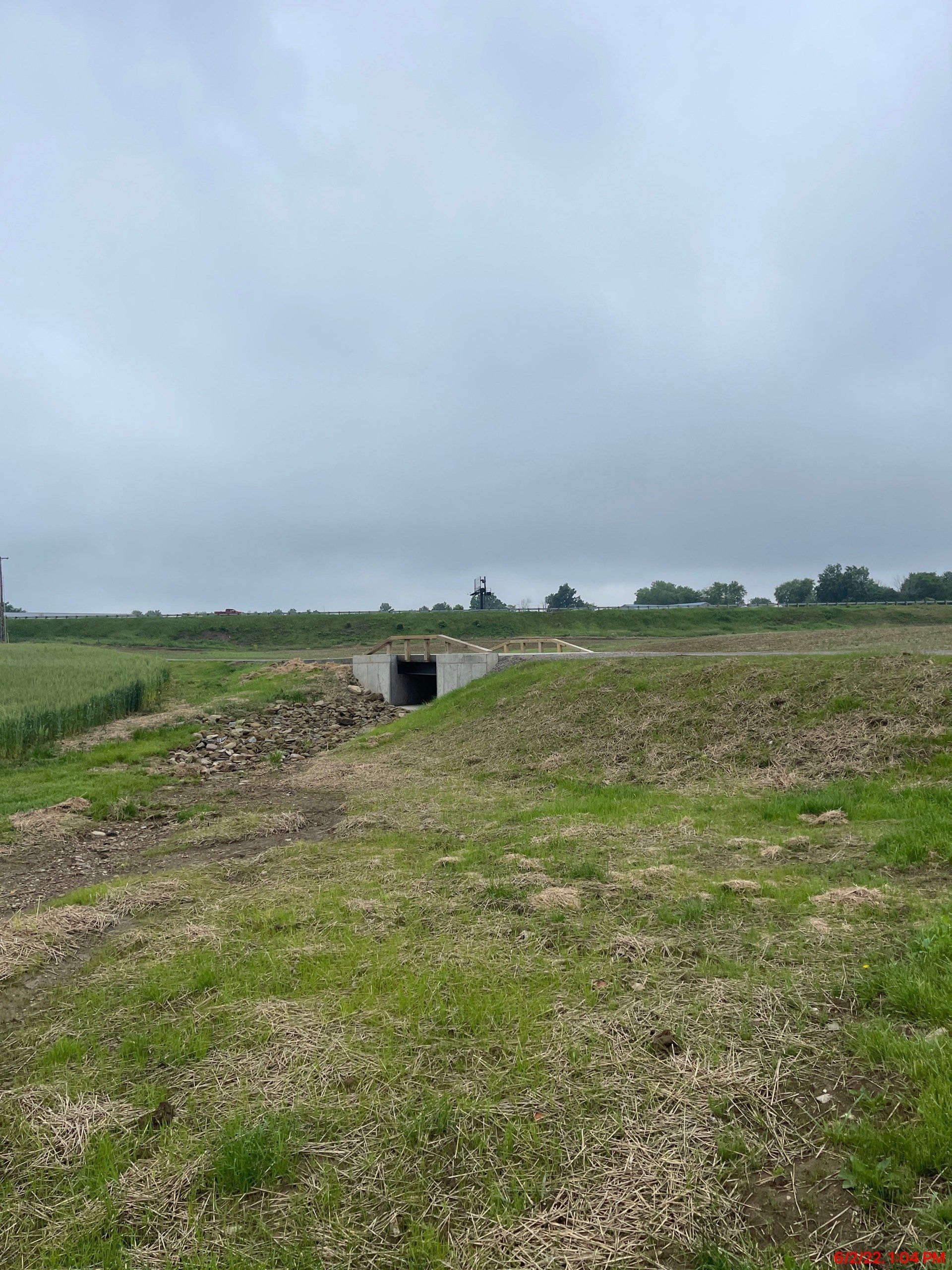 Concrete tunnel entrance in grassy ditch under overcast sky. Farmland in background.