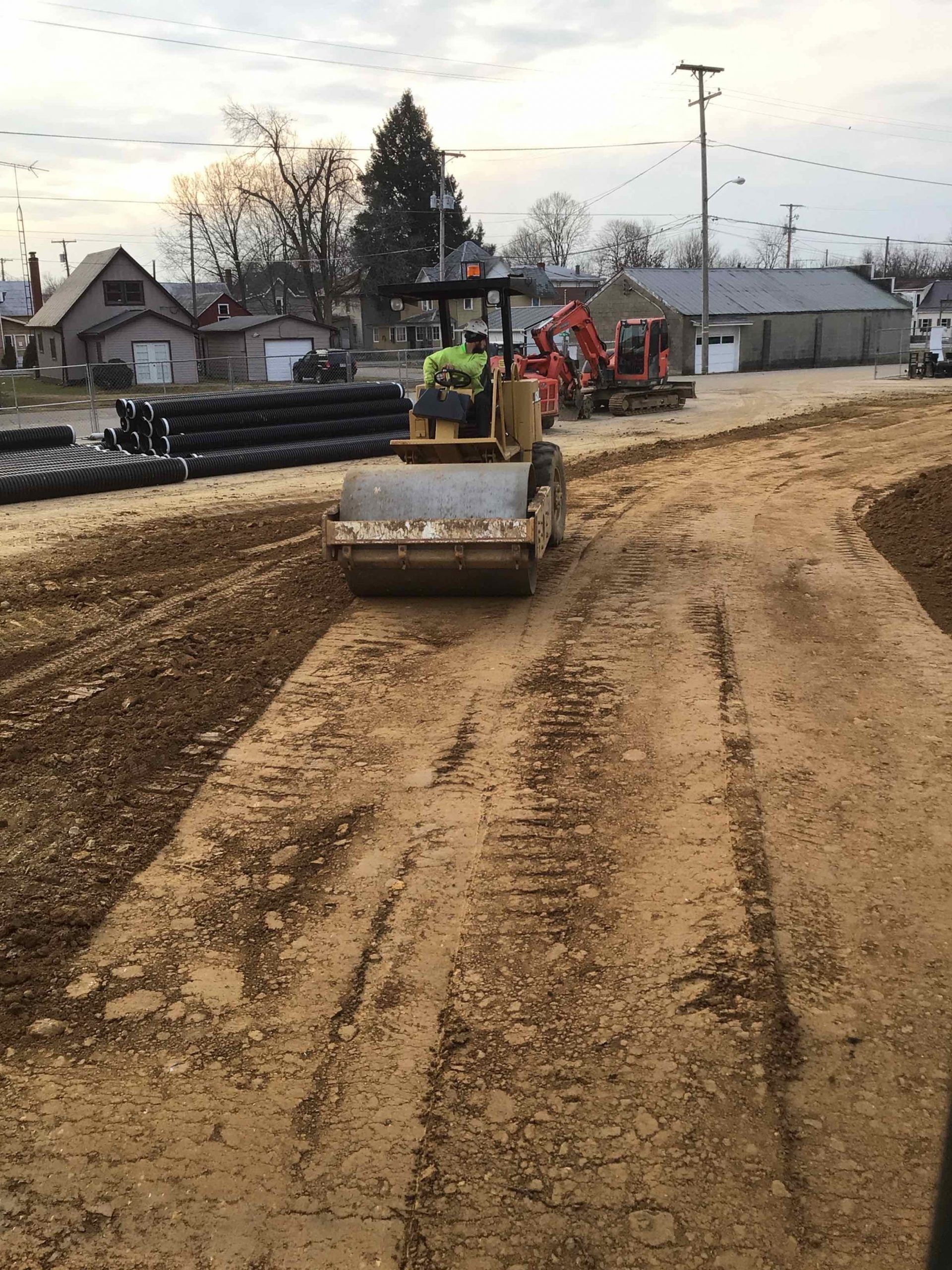 Construction site with a road roller compacting dirt; buildings in the background.