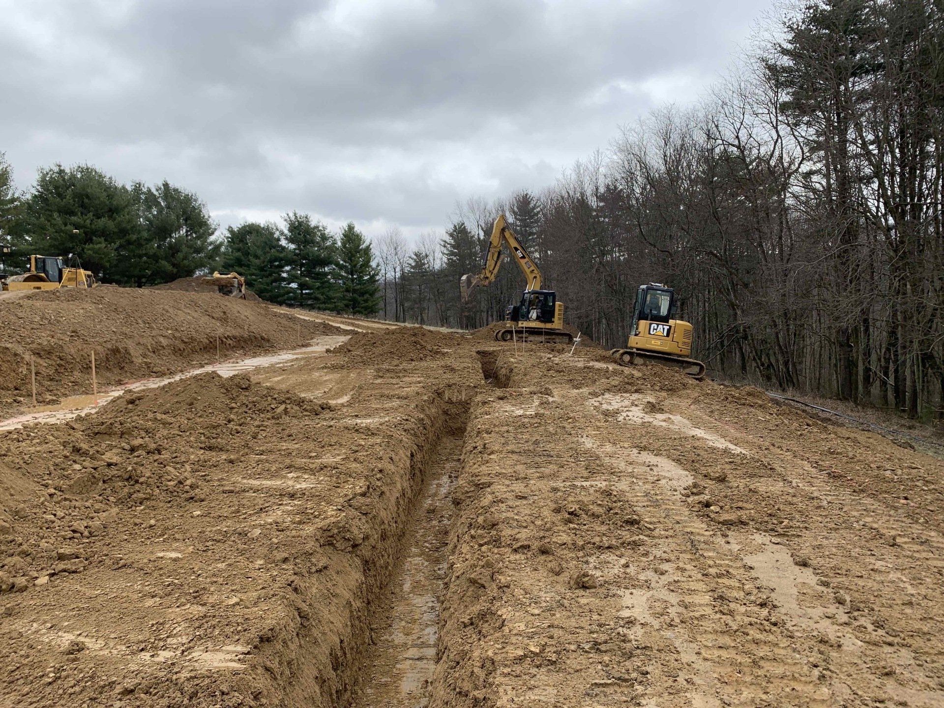 Construction site: Excavators digging trenches in muddy terrain, cloudy sky overhead.