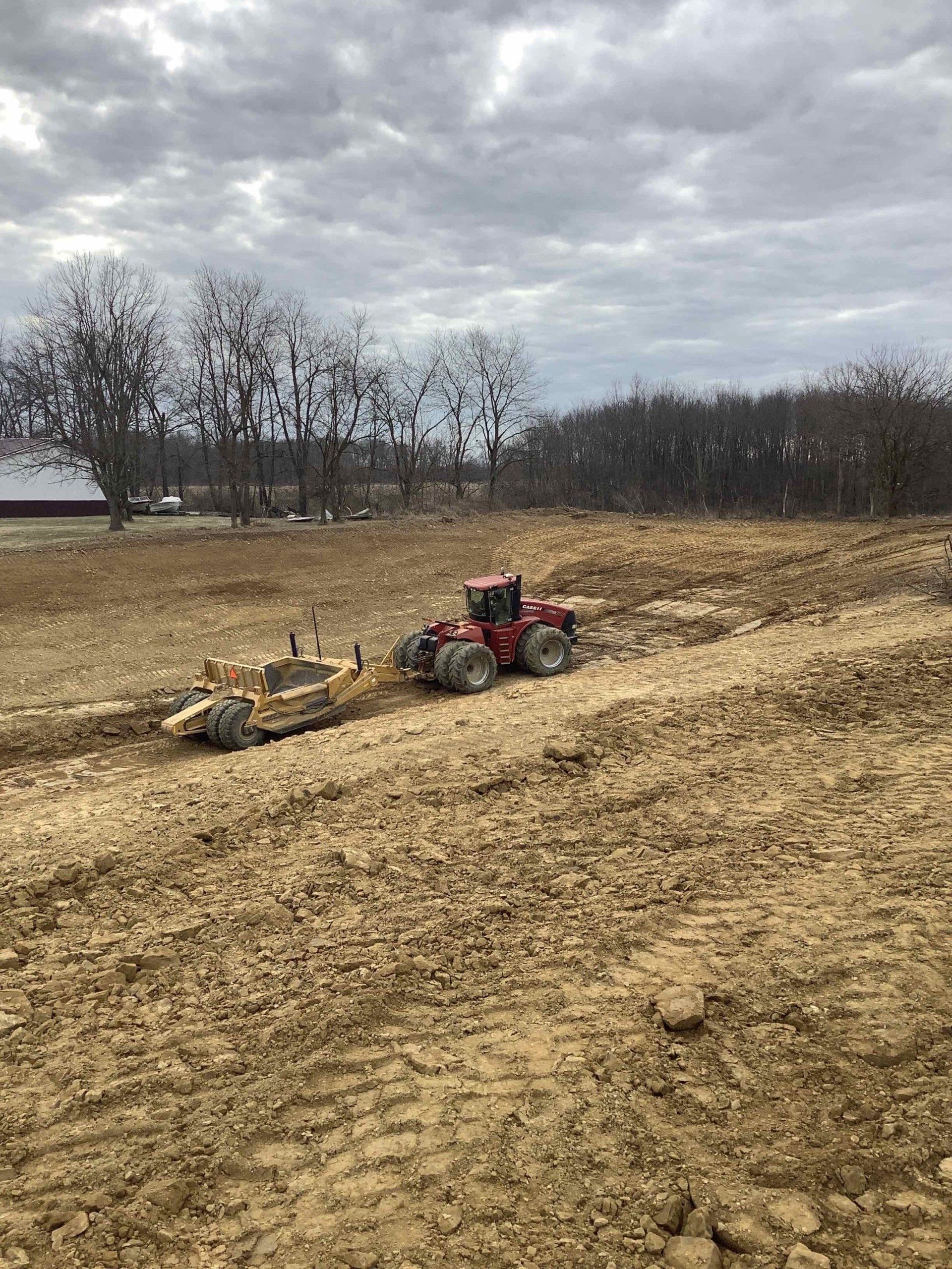 Red tractor pulling a grader on a muddy field under a cloudy sky.