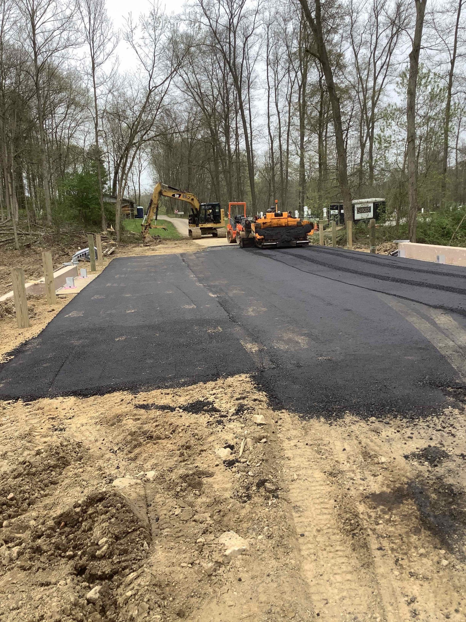 Road paving in progress, featuring asphalt and heavy machinery on a dirt road. Trees in background.