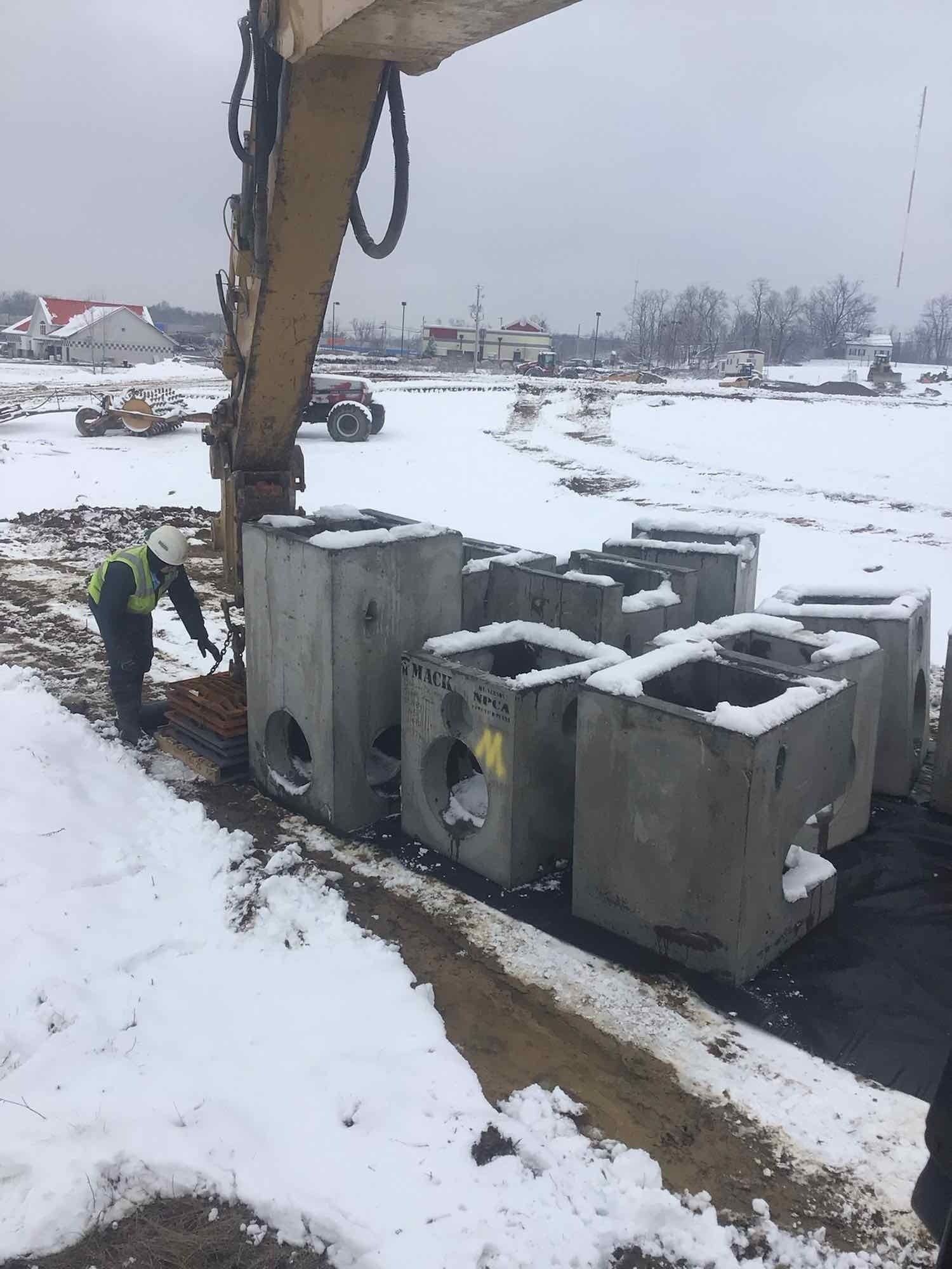 Construction site with excavator placing concrete blocks in snow; worker in safety vest nearby.
