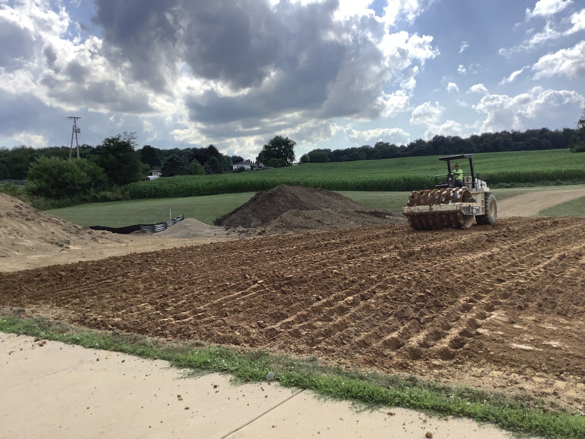 Construction site with a roller compacting dirt; farmland in the background under a cloudy sky.