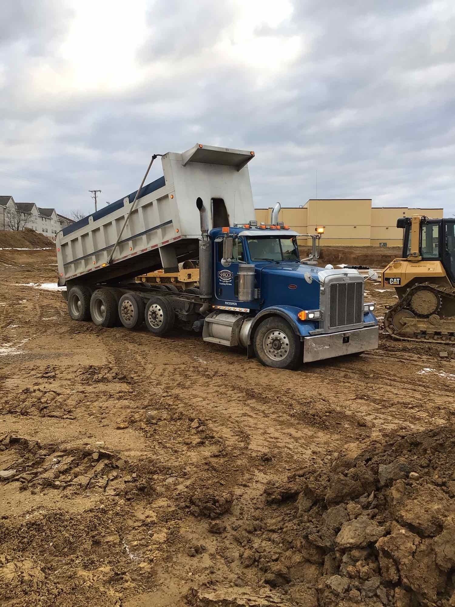 Blue dump truck dumping dirt at a construction site on a cloudy day.