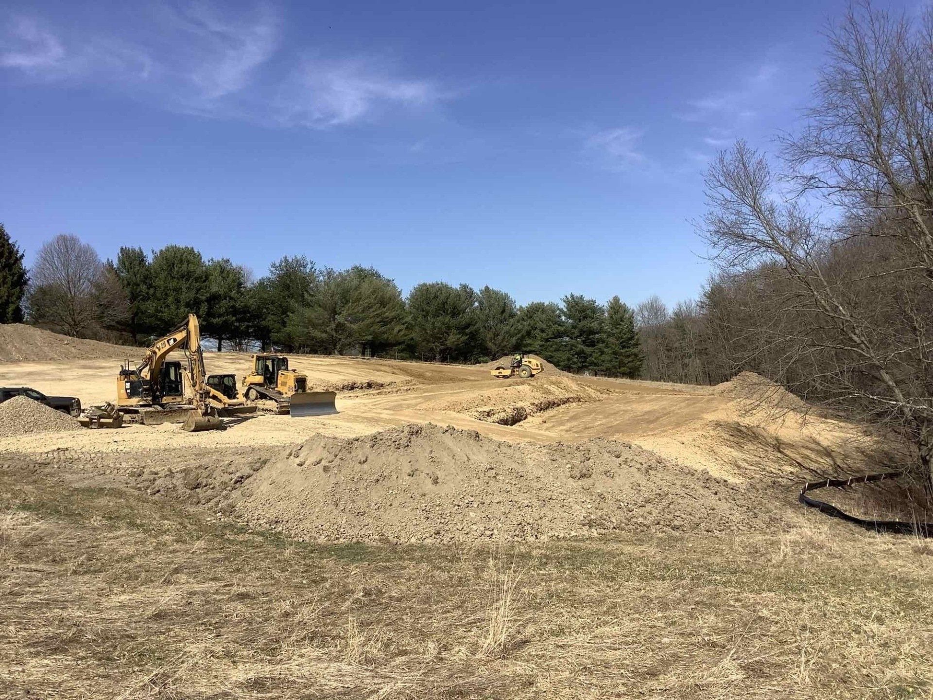 Construction site with bulldozer and dirt piles under a blue sky. Trees line the background.