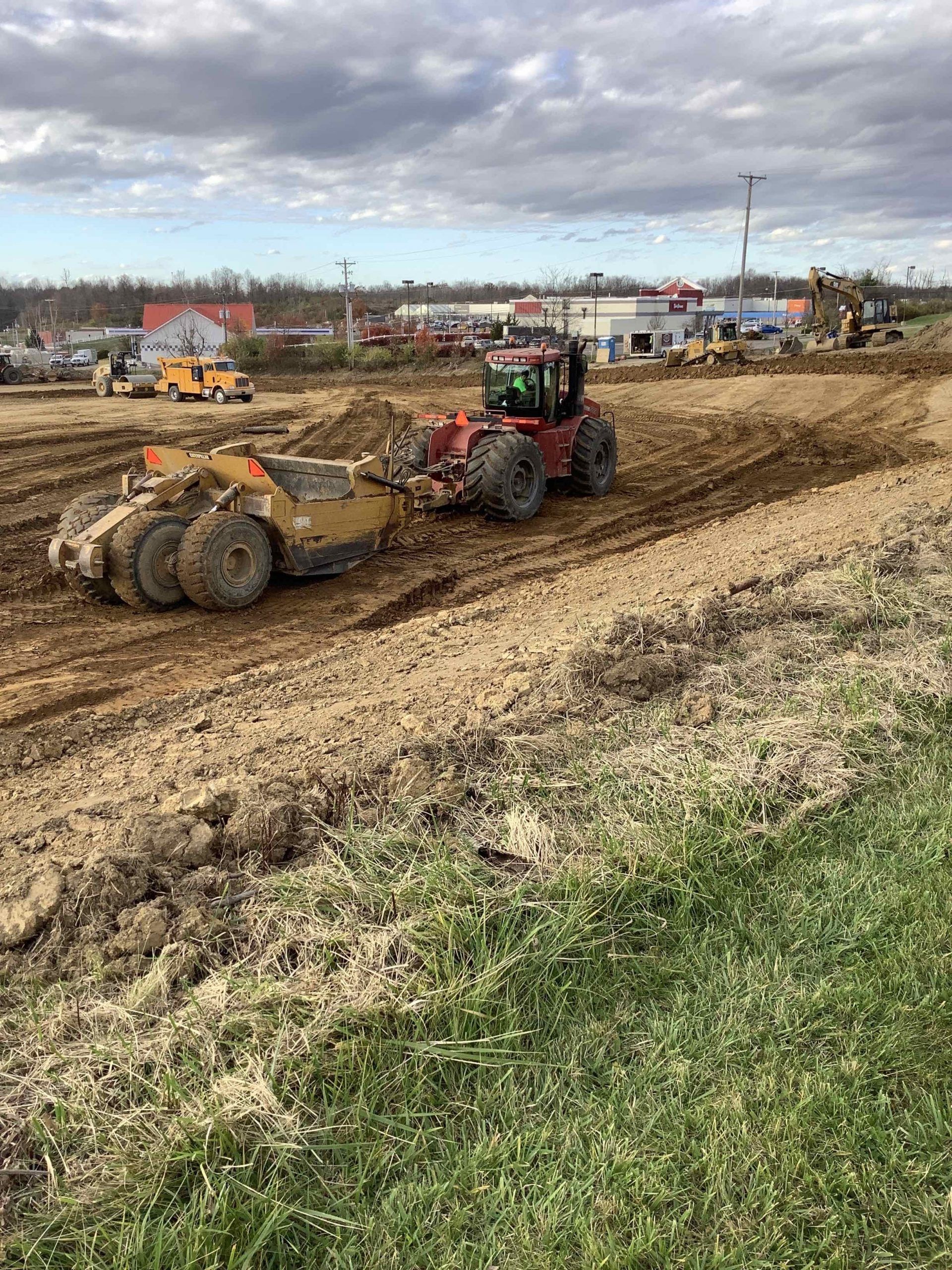 A red tractor pulling a soil scraper on a construction site, cloud-filled sky overhead.