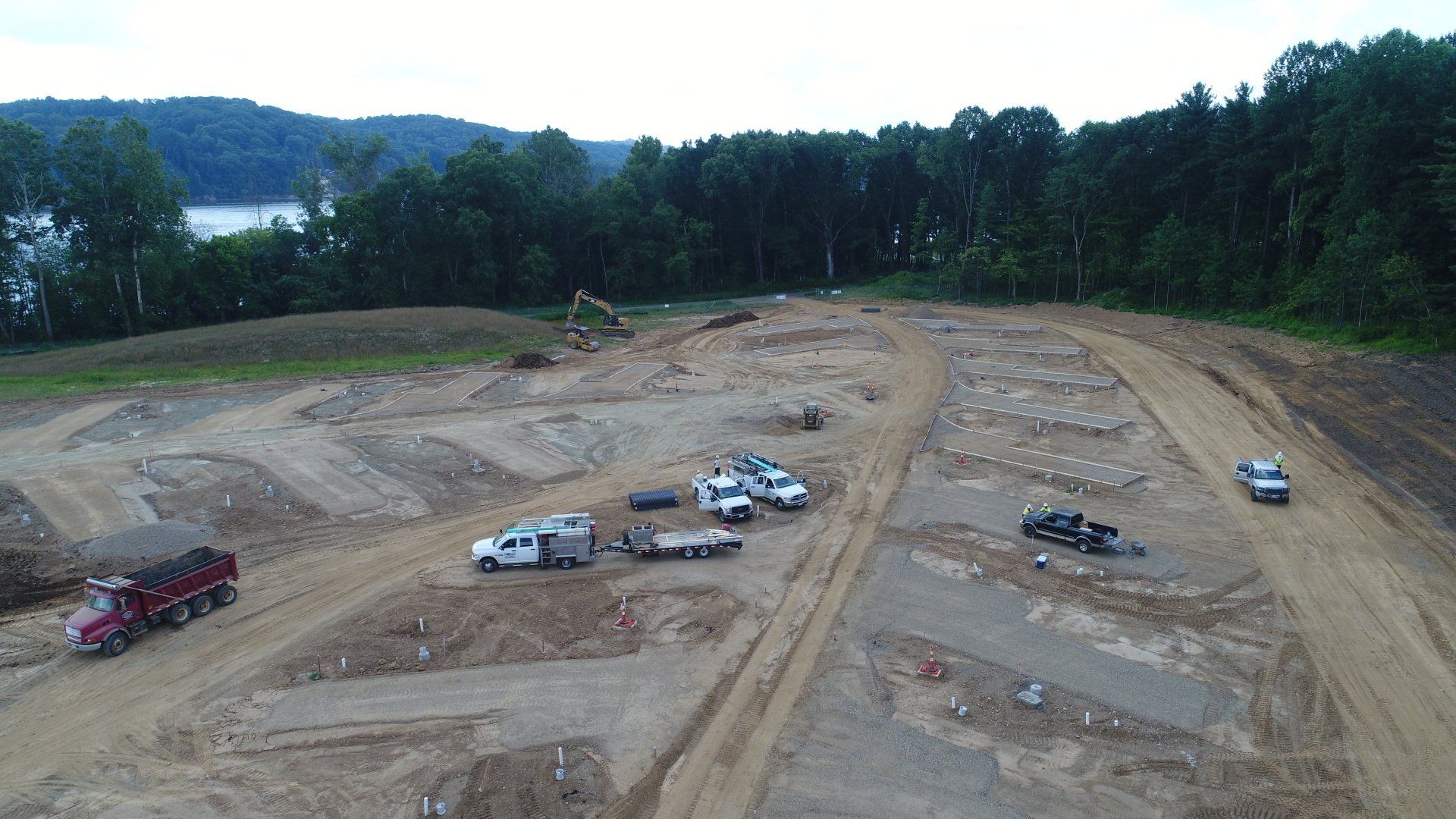 Construction site with trucks, cars, and equipment on dirt; trees and a body of water in the background.