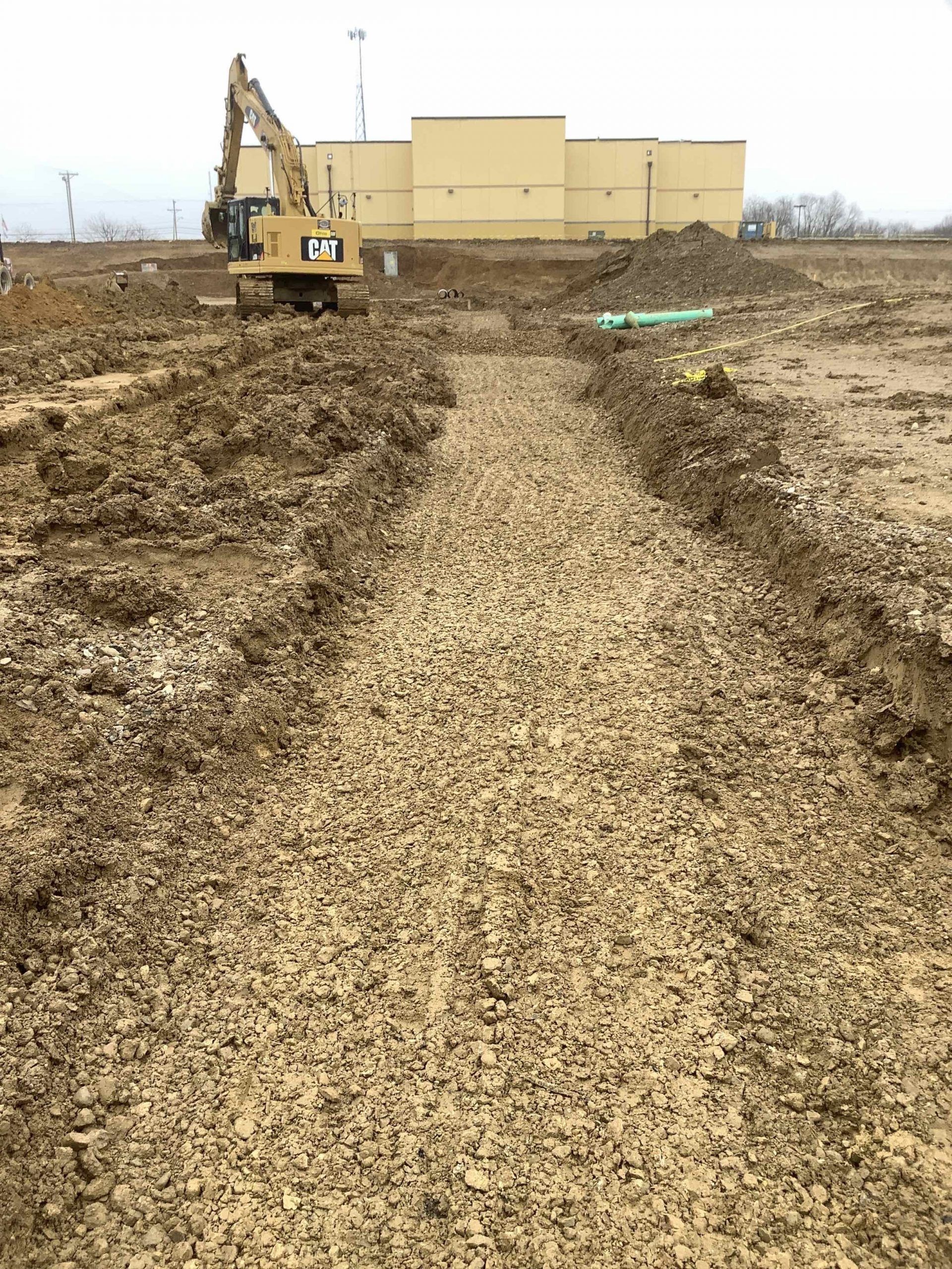 Construction site with a long gravel path; excavator and building in the background.