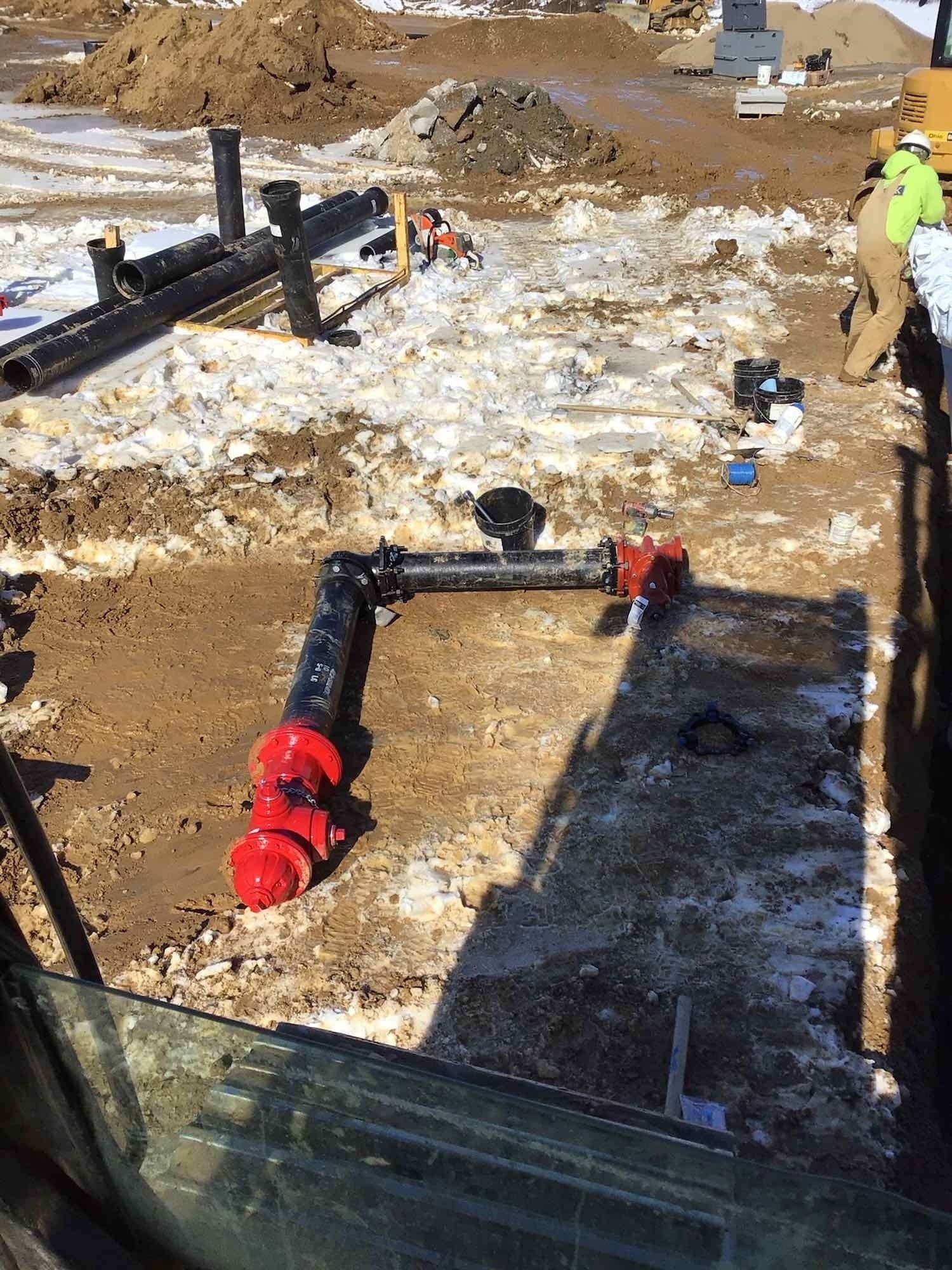 Construction site with black and red pipes in a dirt trench, worker in the background.