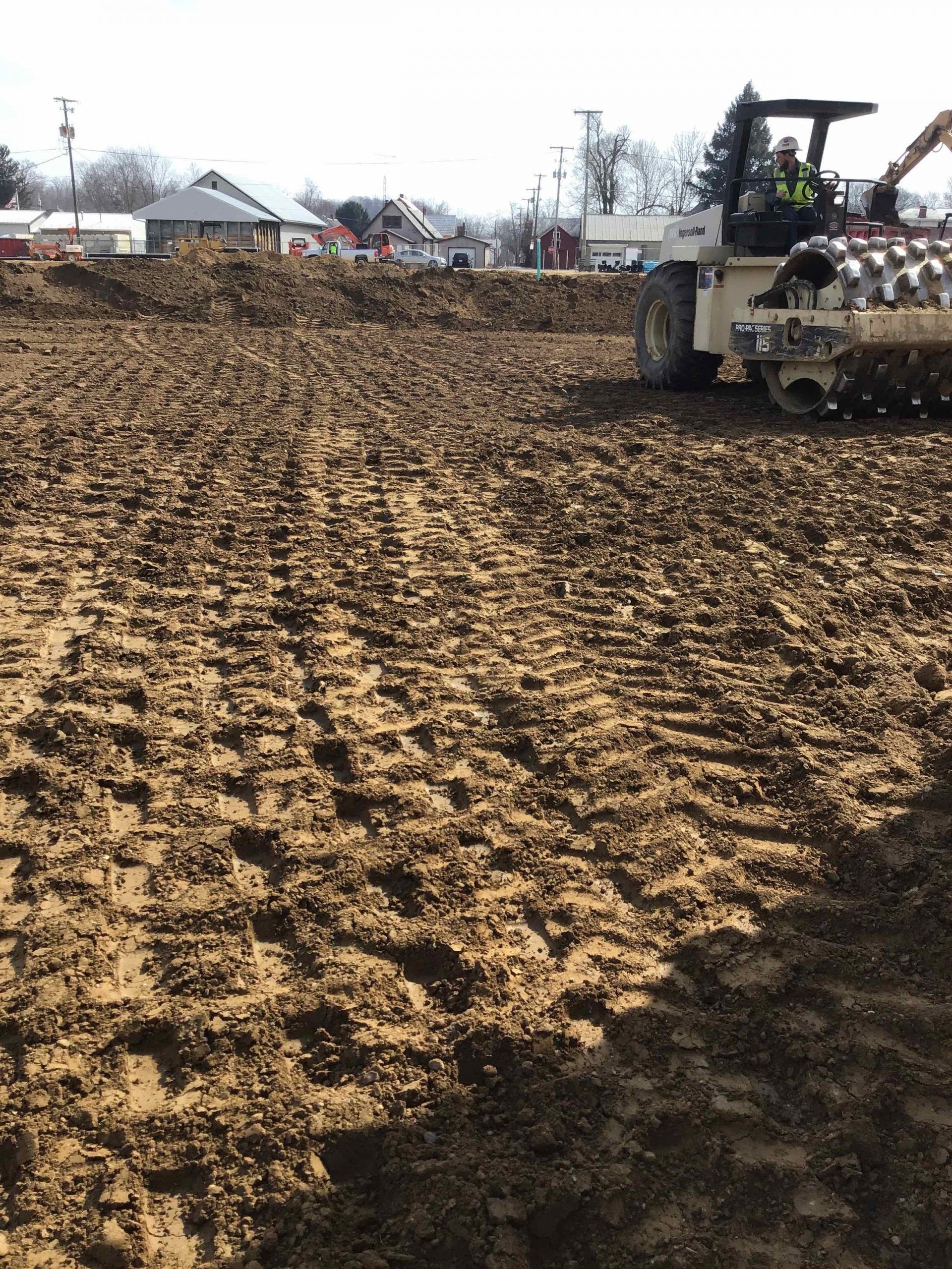A construction site with a large roller compacting brown soil, buildings in the background.