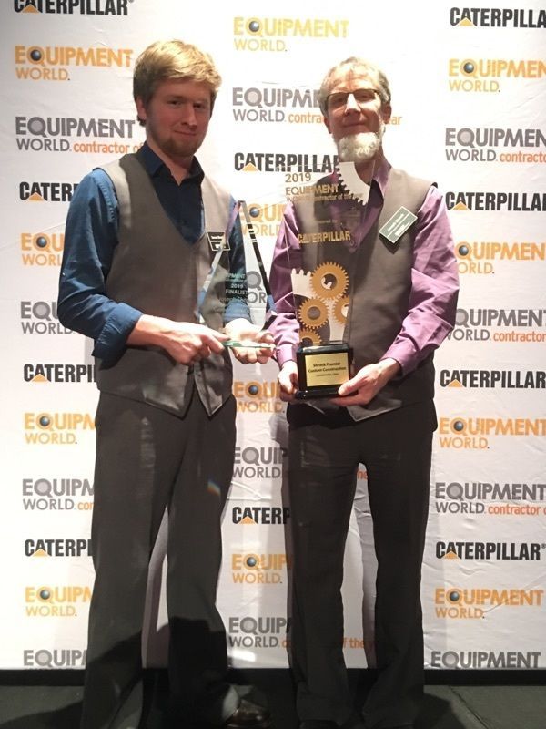Two men holding an award in front of a backdrop with logos. The award features gears.