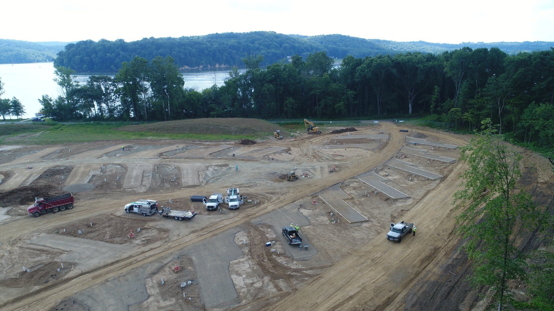 Construction site with heavy machinery and trucks on a hillside overlooking a lake and trees.